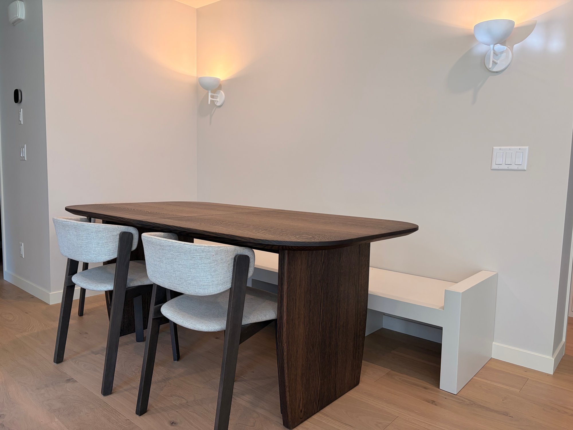 Custom extending dining table in dark-stained white oak with rounded corners and twin panel pedestal bases, paired with a built-in banquette bench in a Calgary dining nook